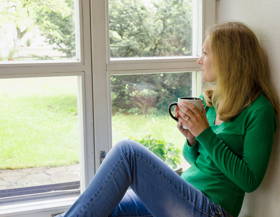 Woman holding a mug with both hands is glancing out a set of white-frame windows.